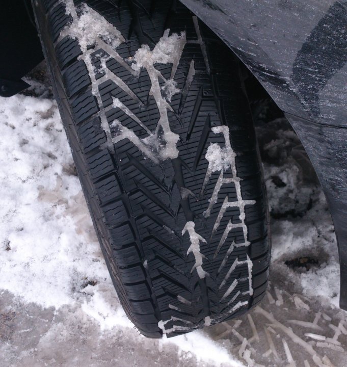 Winter tyres. - Page 1 - Off Road - PistonHeads - The image displays the bottom of a tire, which is covered with a layer of snow and ice. There is a distinct tread pattern, and close to the rim, there are chunks of ice and snow which have been chipped off, revealing a clean area. The perspective of the photo is from the front of a vehicle, and only a part of the vehicle's fender is visible at the bottom edge of the frame. The tire itself appears to be newly purchased or well-maintained.