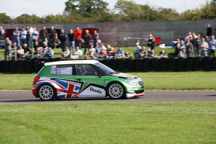 Pistonheads September Castle Rally Day Saturday Coombe - The image showcases a thrilling car race in progress. A small green and white race car is prominently featured in the foreground, driving on a curving track surrounded by lush green grass. The backdrop of the image is filled with a crowd of spectators, all of whom are attentively watching the race. Some spectators are trying to capture the excitement on their cameras. Above the crowd, buses can be seen parked, possibly for the event or for general usage. The overall atmosphere of the image conveys the heightened energy and adrenaline of the motorsport event.