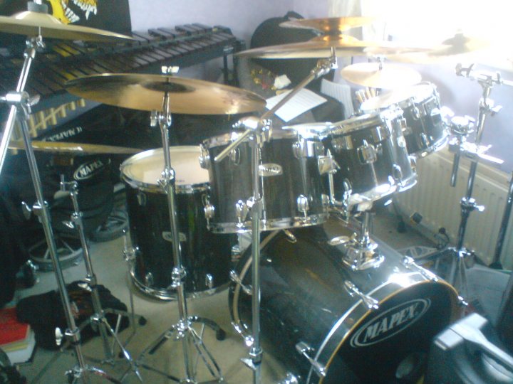 Instruments Pistonheads - The image presents a well-used drum set sitting on a wooden floor in a room with white walls. The drums appear to be in good condition, suggesting frequent practice sessions. The two cymbals and ten drumheads are visible, and there are a few books scattered near the drums, indicating a possible home practice setup. A "Mapex" bass drum with the brand name written in white capital letters is prominent in the middle of the setup. The overall setting appears to be a personal space, likely a home, where music is created and enjoyed.