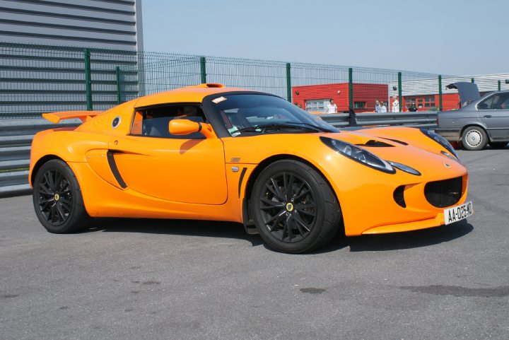 Chrome Orange Exige pictures please !! - Page 1 - Elise/Exige/Europa/340R - PistonHeads - The image captures an outdoor scene with an orange sports car parked on top of what appears to be a parking lot. The car showcases a sleek design with distinctive features that include a distinctive rear wing, large wheel arches, and a front fascia face with the brand's logo prominently visible. In the background, there are buildings and a red vehicle, which could be a truck or a car, is partially visible. The lighting suggests it might be daytime, as the sky is clear and bright.