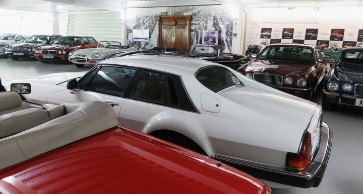 Pistonheads - The image shows an interior view of a car showroom with various cars on display. In the foreground, there are two vintage cars, one with an open hood and the other appearing to be a classic convertible. These cars are situated in the center of the room and are prominently displayed. Behind these vehicles, there is another row of cars that are partially visible. The background features a high ceiling and white walls, which suggests an institutional or corporate setting. There is no visible text or branding within the image.