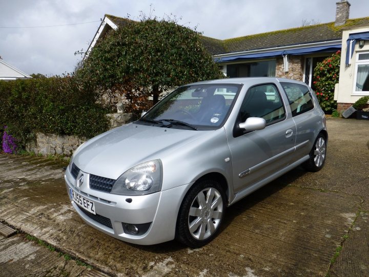 My new PH's car! - Page 1 - South West - PistonHeads - The image shows a small, silver-colored hatchback parked on a paved driveway. The car's body has some residue or dirt on it, suggesting that it has been used. In the background, there is a house with a shingled roof and a small hedge. The cloudy sky hints at an overcast day. The overall scene appears to be a typical residential setting.