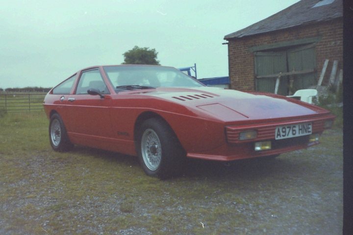 Pistonheads - The image features a vintage red sports car parked on a grassy area. The car, with a distinctive angle, stands out prominently in the scene. In the background, there's a classic brick building with a sloped roof, and a wooden lean-to structure is also visible, adding to the nostalgic atmosphere. The photograph has a vintage filter, which enhances the old-world charm of the setting. This composition suggests a tranquil countryside setting featuring notable symbols of the past.