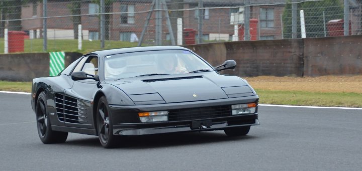 testarossa thread - Page 1 - Ferrari Classics - PistonHeads - The image shows a dark-colored sports car in motion on what appears to be a track. The vehicle has a sleek and streamlined design typical of high-performance cars. Its position indicates that it's in a left-hand turn. In the background, there is a blurred landscape that appears to be a course with fencing along the sides. The setting suggests that this could be a race or a private driving event. The car's design and the circumstances hint at a late model, possibly mid to late 20th century.