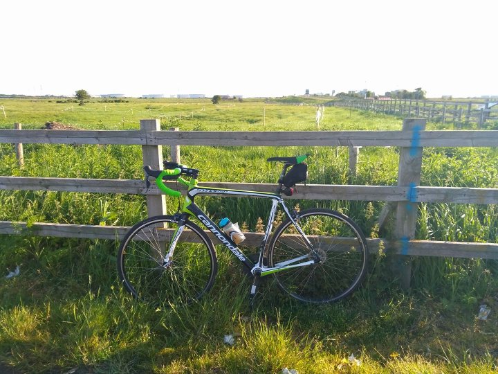 Cannondale Synapse or Supersix? - Page 1 - Pedal Powered - PistonHeads - The image presents a tranquil outdoor scene featuring a **black bicycle** leaning against a light gray wooden fence. The bike is equipped with a **black bag**, perhaps for a long journey, and is scattered with various accessories. The fence, weathered and rustic, stands sturdy against a lush green field that stretches out behind it, eventually leading the eye towards a **line of trees** beyond. The overall atmosphere exudes a sense of calm and solitude, with the bike standing as a touchstone amidst the natural beauty. The photo captures the lively and unexplored world possible on two wheels.