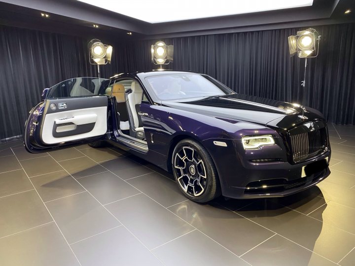 The image shows a luxurious vehicle, which appears to be a customized Rolls Royce car. The car is parked indoors and has its doors open. It features a distinctive paint scheme, with one side painted in a lighter color than the other. There are no visible people or texts that provide additional context about the location or event. The interior of the car is partially visible through the open door. The setting suggests a private display or showroom, indicated by the presence of stage lighting and curtains.