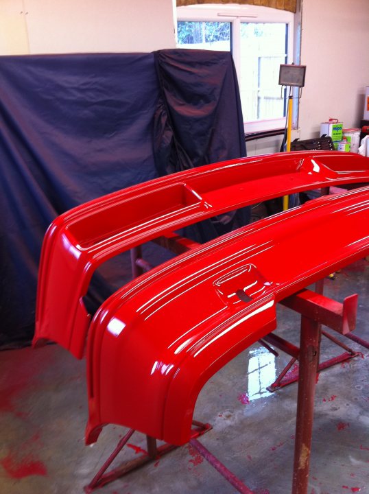 Fiesta RS Turbo Restoration - Page 6 - Ford - PistonHeads - The image depicts a red bumper being painted in a garage, with the car's reflection visible on the concrete floor. The bumper sits on a metal stand on the right side, ready for paint application or touch-up. In the background, there is a window with a partially visible board and a door with a view slit. The setting suggests a workspace for automotive restoration or customization. The paint has already been applied to a part of the bumper, which indicates it might be a part of the vehicle that has seen some wear and is being refreshed.