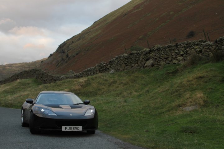 black evora spotted - Page 1 - Evora - PistonHeads - The image features a modern sports car navigating a winding country road. The car is black, with a sleek design, and is traveling away from the viewer. The scenery is picturesque, with a dry stone wall alongside the road, which twists from the foreground into the distance. The terrain is hilly, with verdant grass and a rocky hillside that's characteristic of a rural, hilly setting. The sky is overcast, suggesting a cool, clear day.