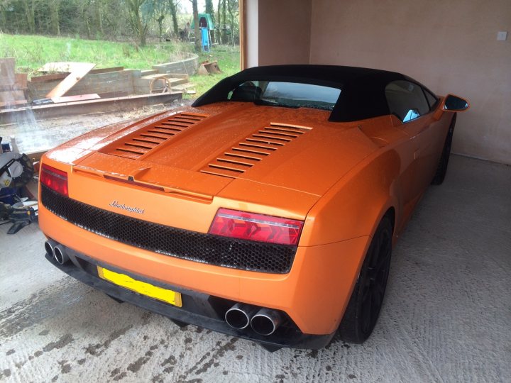 More pics of my LP560 - Page 1 - Gallardo/Huracan - PistonHeads - The image shows an orange sports car parked in a driveway. There is a person visible near the car but only their lower body is seen. The car appears to be a convertible model with its top up, and it has a pointy rear with two red taillights. The background suggests the setting is outdoors, with some structure visible and a glimpse of greenery.