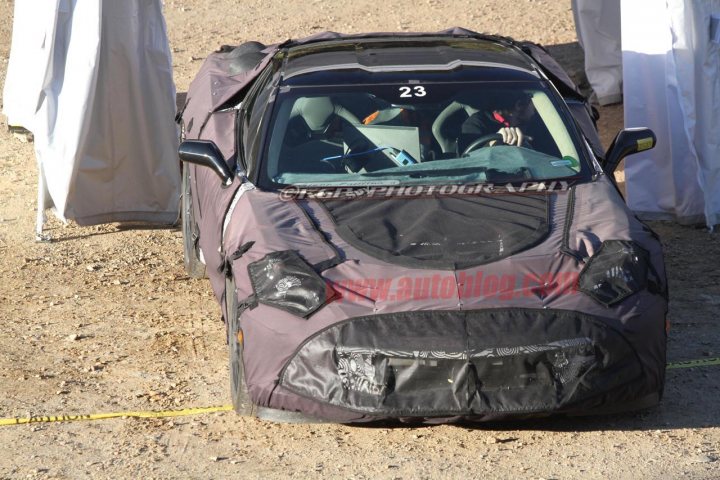 C7  Bucket seats and glimpse of the new steering wheel - Page 1 - Corvettes - PistonHeads - The image shows a small, two-door convertible car that has been completely covered with a thick black material, making the car look finished and without bumps or scratches. The car is parked on a dirt surface with a yellow measuring tape coiled loosely on the ground beside it, suggesting that it might be part of a project or exhibit. In the background, we can see outlines of figures that appear to be part of the display, but they are not very visible. The atmosphere of the image is industrial or workshop-like, suggesting that this car is being showcased or prepared in a specialized setting. The car appears to be in excellent condition, which is accentuated by the smooth black material wrapping it.