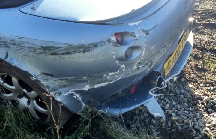 Crashed Tam, advice needed on how to go about repair.  - Page 1 - General TVR Stuff & Gossip - PistonHeads - This image features the side view of a grey car. The car's front side is damaged, with a notable dent in the quarter panel and a portion of the fender missing, revealing the underlying metal components. The vehicle is parked on what appears to be a patch of grass or a small plant. The surrounding area suggests a rural or remote location, with the ground appearing rocky and uneven. There is also visible damage to the nearby trees, indicating a possible collision or impact. Due to the loss of context and the focus on the vehicle, no textual or human subjects are present in this image. The image provides a close-up view of the damage, emphasizing the impact of the incident.