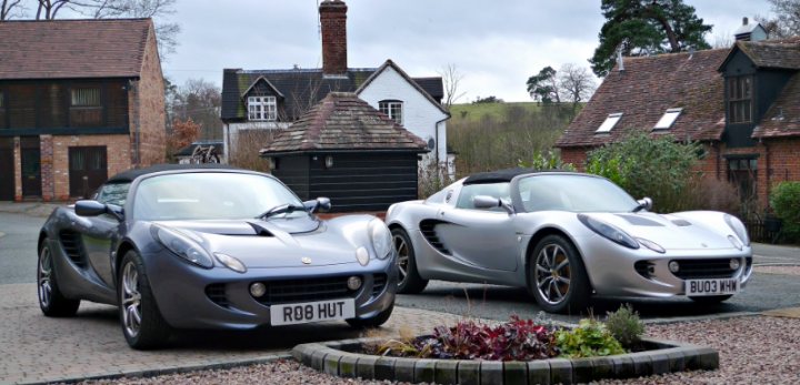 Headlamps on an 111R '06. - Page 1 - Elise/Exige/Europa/340R - PistonHeads - The image features a serene outdoor scene with two sports cars parked on a brick driveway. The car on the left is predominantly dark with slightly lighter detailing. The car on the right has a metallic finish, creating a sleek and modern appearance. Both cars are low to the ground with long hoods and short rear decks. The setting includes a quaint, traditional-style house with a thatched roof behind the car on the left. The driveway leads to a cul-de-sac lined with similar houses, one of which has a visible presence of nature, as there are leaves scattered on the curb. The sky above is dark and cloudy, suggesting either an overcast day or early morning. There is no visible human activity in the image. The overall aesthetic of the image is calm and orderly, with an emphasis on the beauty and sleekness of the vehicles.