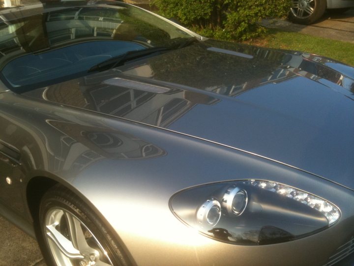 Pistonheads Slinc - The image shows a close-up view of the front portion of a silver luxury car. The car's metallic sheen is highly visible, reflecting the surrounding environment, which includes tree branches and a building. The headlight of the car is prominent, showing off its high-end design with intricate detailing. Inside the reflection on the car's hood, it's possible to discern the shapes of an umbrella and another object on a nearby table, suggesting that the car is parked near a dining or working area. There is also a reflection of a car wheel in the image. The overall scene suggests a well-maintained, parked vehicle in an urban or semi-urban setting.