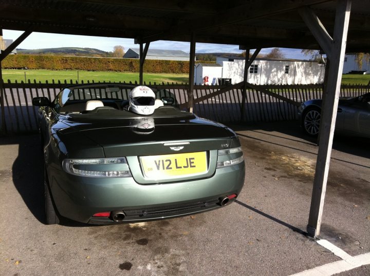 Goodwood Aston day - Page 1 - Aston Martin - PistonHeads - The image depicts a scene in a parking lot. A green Aston Martin DB7 luxury sports car is parked in a space, showcasing a sleek and elegant design. The car's license plate is clearly visible, indicating "V12 LJE" as the registration. On the hood of the car, there is a white helmet with markings, suggesting that the driver may be involved in racing or an event that requires such protective gear. The ground appears to be wet, as indicated by the reflections on the asphalt. The background features a chain-link fence and a distant building, providing context for the location of the parking lot.