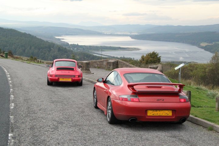 Highlands - Page 71 - Roads - PistonHeads - Two vibrant red sports cars are parked along the side of a road, set against a picturesque backdrop of a body of water and distant mountains under a clear sky.