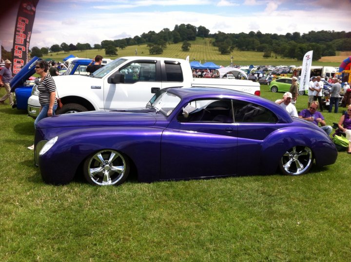 A car is parked on the grass with a dog - Pistonheads - This image captures a vibrant car show scene. The main focus is a stunning purple vintage car, which is parked along with several other vintage cars on a grassy area. A white pickup truck is also visible in the background. Above them, several tents and flags are set up, suggesting that the event might be covered or inclusive of some form of activities or demonstrations. The crowd, scattered in the background, is likely admiring the beautifully restored vehicles.