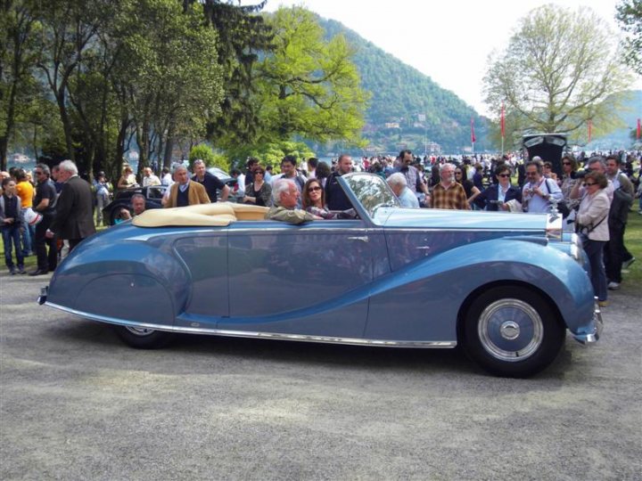 Villa Concoursporn Pistonheads Deste - The image captures a lively outdoor event. In the foreground, a crowd of people is gathered around a vintage blue convertible car. The car, with its convertible top up, presents a classic design that draws attention. It is parked by the side of the road, with a glove on the passenger seat. The crowd appears interested in the car, indicating that it might be of particular interest or showcased as part of the event. The setting suggests a sunny day with good weather, which is likely conducive to such outdoor exhibits.