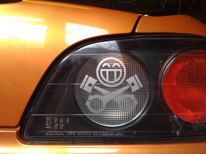 Pistonheads Rice Burner - The image captures an upfront view of the tail light assembly of an orange car, with a distinctive feature being a logo affixed to the licence plate area. The logo is a smiley face, adding a touch of personality and charm to the vehicle. The shiny surface of the car reflects some sounds, indicating a recent drive. The license plate itself shows various characters, but the text is not clear from this angle. The reflection suggests a sleek design for the car.