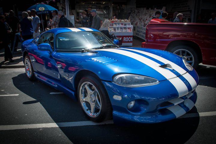 A car that is sitting in the street - Pistonheads - The image showcases a striking blue sports car, parked in an outdoor lot. The vehicle's sleek design is accentuated by a bold white stripe running the length of its hood. The car's front is characterized by a large, chrome grill and alloy wheels, hinting at its high-performance capabilities. A black privacy tint blankets the windows, while a shiny, blue paint job glistens under the sunlight. The car is the main focus of the image, set against a backdrop of a sidewalk lined with pedestrians and an open stall.