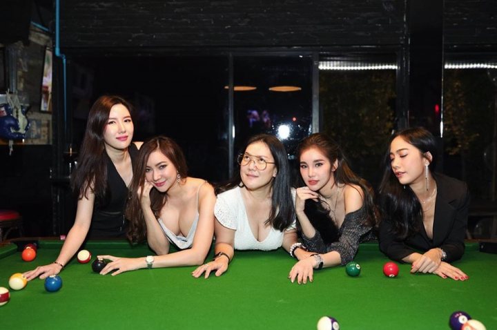 A group of women standing next to each other - The image is a photograph of a group of women posing for the camera in what appears to be a billiards room or pool hall. There are five individuals visible, with four standing and one seated in front of them. All five are dressed in smart casual attire and are smiling at the camera. They are gathered around a pool table that has several cue balls on it. The room has a dark ambiance with a mirror reflecting part of the scene behind them. There is no visible text within the image.