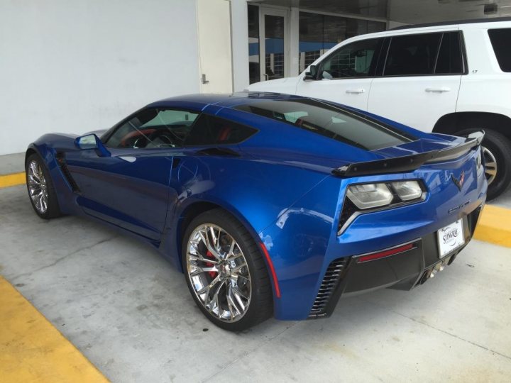 Daytona and Stingray Chevrolet (Z06). - Page 1 - Corvettes - PistonHeads - The image features a vibrant blue Chevrolet Corvette parked in a parking lot next to a white SUV. The Corvette is parked slightly off-center to the left, facing towards the right side of the picture. The car's aerodynamic design is accentuated by a prominent rear spoiler and a clean, shiny finish to its exterior. Behind the sports car, you can see another vehicle, suggesting the lot is busy. The parked vehicles are under clear skies, contributing to a well-lit, bright day in the image. There is no visible text in this image.