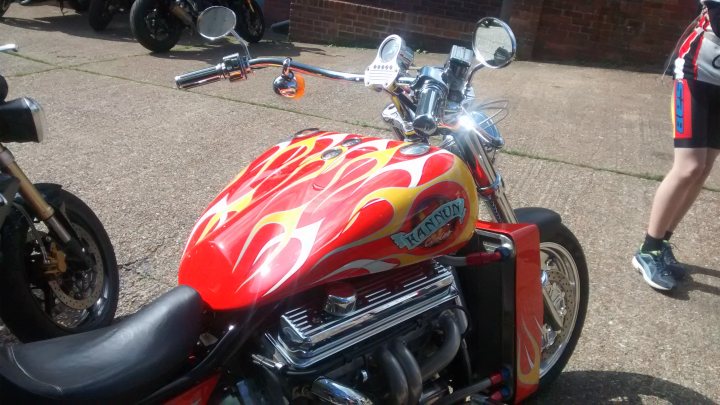 The "Photos From Today's Ride" thread... - Page 168 - Pedal Powered - PistonHeads - The image showcases a striking red motorcycle with an elongated gas tank. The motorcycle is adorned with intricate flame designs on its gas tank, adding to its distinctive look. It's parked on a street with several motorcycles on either side in the background, suggesting a gathering or event of bikers. The motorcycle's black seat contrasts with its red body, completing the eye-catching color scheme.