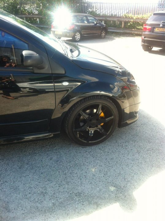 C2 VTR- brakes - Page 2 - French Bred - PistonHeads - The image shows the side of a black sedan parked on a concrete surface. The photo captures the car's rims, reflecting light, and is taken under bright sunlight. There are blurred reflections of the photographer and the outside surroundings visible in the glass surface of the vehicle. There appears to be a shadow adding depth to the image, cast by the car, giving a sense of direction.