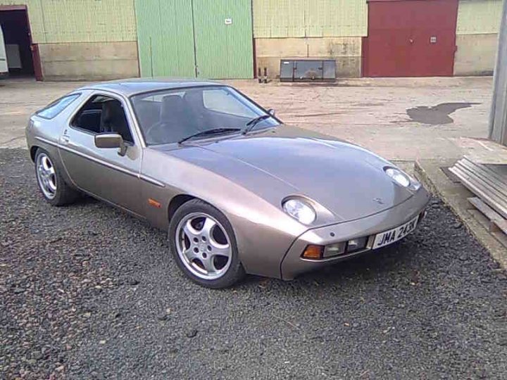 Porsche 928S - Page 1 - Readers' Cars - PistonHeads - In the image, we see an old-style car parked in a gravel lot within an industrial setting. The car has some grayish-silver hue and features a unique twin-roundel design, indicative of the Alpine model, which is also confirmed by the registration plate. The car's position next to a small structure and door suggests it might be part of a compound or collection. The backdrop includes buildings and additional doors, reinforcing the industrial context.