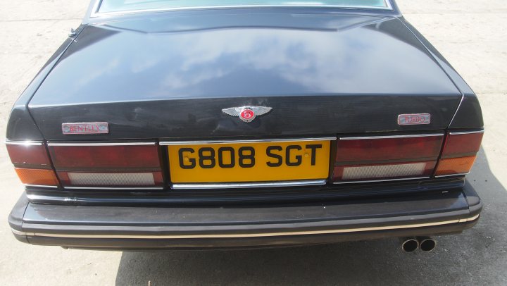 Have we been foolish? Yes but we're all grinning from ear to - Page 1 - Rolls Royce & Bentley - PistonHeads - The image features the back of a black car parked on a pavement. The car's registration plate reads "G808 SGT." Above the license plate, there's a small, square emblem. The car has dual round tail lights, a common style for some vintage or classic car models, and there's an exhaust pipe visible. The sky in the background is partly cloudy, and the ambiance of the image suggests a calm or rural setting.