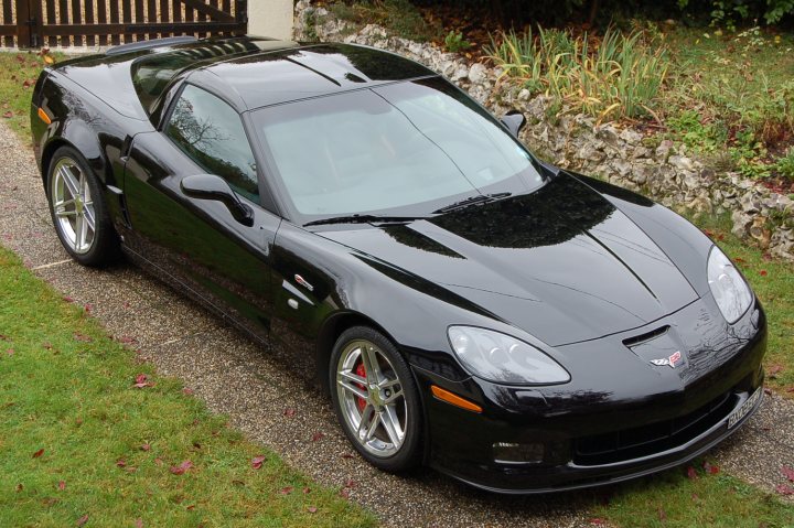 Pistonheads - The image displays a black sportscar parked on a gravel driveway. The car has a sleek design with a prominent package at the rear. The vehicle is angular and features large wheels and a low stance. It also includes air vents behind the doors, which is typical for sports cars to help in cooling the engine. The setting appears to be a residential area with grass around the driveway and foliage in the background.