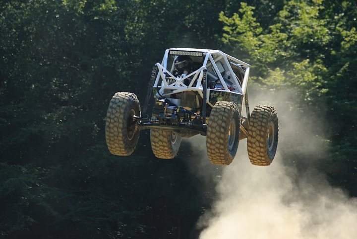 Flying 4x4s - Page 1 - Off Road - PistonHeads - The image showcases an exciting moment in what appears to be an off-road race. A Monster truck, bearing the number 46, is captured mid-air above a dirt track, flipping over its side in a dynamic display. The truck is predominantly white, contrasting with the darker tones of its four large tires and the background. The scene is set in a forested area, with trees lining the track and the sky gleaming through the canopy, adding to the thrilling atmosphere. The image encapsulates the adrenaline-driven spirit of such truck racing events.