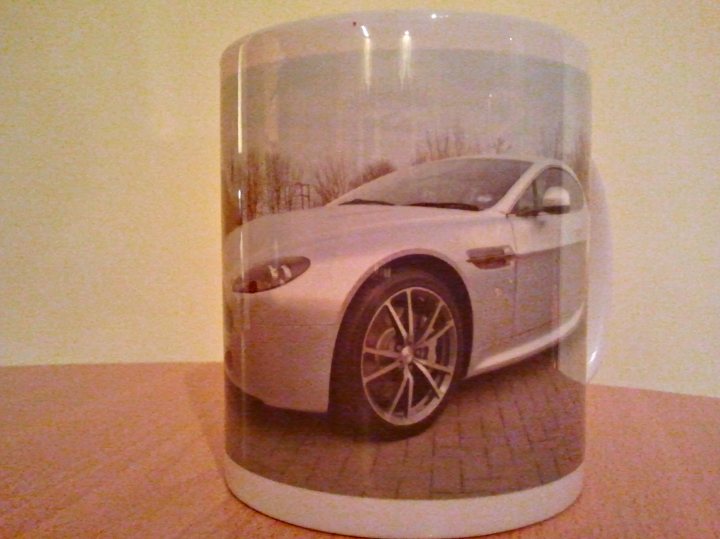 A right Mug! - Page 1 - Aston Martin - PistonHeads - The image features a white mug with a printed photograph of a silver sports car. The mug is placed on a wooden surface, potentially a table. The car, the central object in the photograph, has a convertible design and is presented from a side angle. Its wheels are clearly visible and it appears to be parked on a brick pavement. The photo on the mug conveys a sense of luxury and fine automotive design.