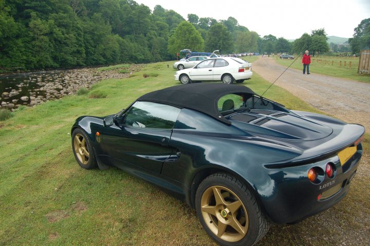 The big Elise/Exige picture thread - Page 6 - Elise/Exige/Europa/340R - PistonHeads - The image shows a black Lotus car with a convertible top, parked on a grassy patch near a country road. The car's metallic automotive finish contrasts with the natural greenery surrounding it, including a riverbank and trees in the background. In the distance, there's a group of people gathered near a roadway, suggesting some event or gathering might be happening. The sky overhead is clear and blue. The car is positioned with the back towards the camera, allowing us to appreciate the design of the car from its side.