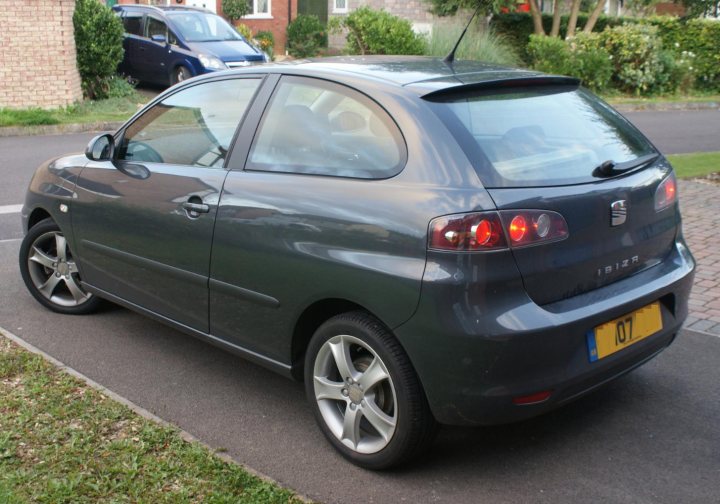 Pistonheads Seat Sport Whip Ibiza - The image showcases a dark-colored car parked on a residential street. The vehicle is a hatchback model, characterized by a sloped rear window and a spare tire mounted on the back. There is an object next to the car, seemingly another motor vehicle, but it is not the main focus of the image. The house behind the car features a red door, adding a pop of color to the scene. The overall image portrays an everyday suburban scene.