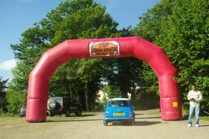 Pistonheads London Brighton - The image features a vibrant scene, likely taken at a parade or car show. A small blue car is driving under a large red inflatable arch with the text "STOP LONDON MINI Burn It" printed on it, suggesting it's part of a themed event celebrating a brand of mini cars. In the background, there are two people standing, one of whom is holding a leash, indicating a casual and relaxed atmosphere. On the left side of the image, there's a black SUV parked, adding to the event-like setting. The trees and sky in the background give this cheerful event an outdoor ambiance.