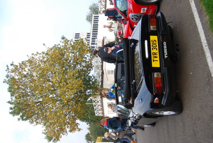 Hi from a newbie 3000S owner - Page 1 - Classics - PistonHeads - In this image, a convoy of small cars is traveling down a street that stretches into the distance. The back of a black sports car is prominently displayed in the foreground, and a person with a camera is following closely behind. The cars are closely packed together, implying they are part of a group or convoy. The street is lined with trees and buildings, and pedestrians are visible at the curb, watching the passing vehicles.