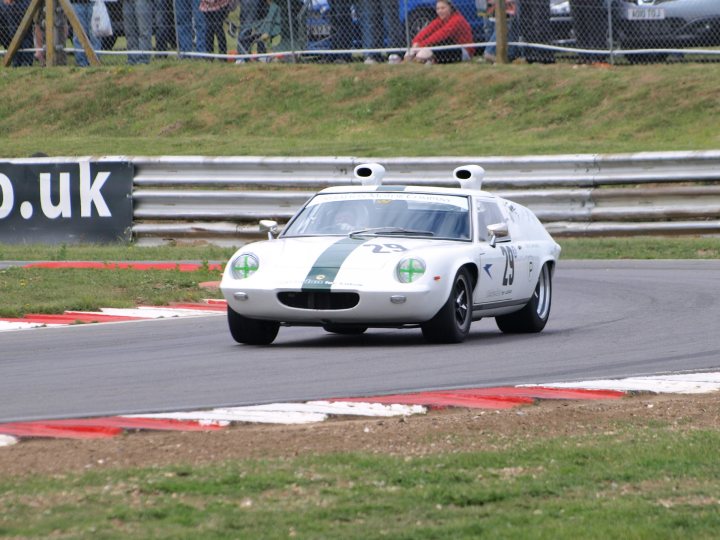 Pistonheads Snetterton - This image captures a dynamic scene at a race track. The main focus is a white race car, featuring distinctive green and blue accents, speeding around a corner. The car's sleek design is complemented by its black wheels and a rear spoiler. The track itself is lined with red and white striped barriers, providing a vibrant contrast to the verdant green of the grass around the track. Although the spectators are not directly visible in the frame, their presence is implied by the fence running along the upper edge of the image, suggesting they are watching the thrilling race unfold.