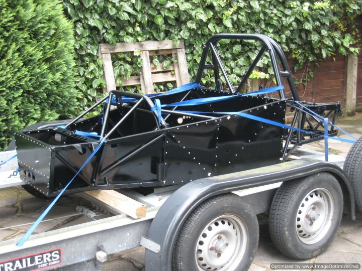 A large truck is parked in a field - Pistonheads - The image shows an old racing car on a trailer, with a green hedge in the background. The car is predominantly black with blue straps tied around it. There appears to be some financial aid information at the bottom. The trailer and car are casting a shadow on the ground. The overall scene suggests a preparation for a classic racing car display or event.