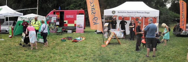 Paws in the Park Today - Page 1 - Thames Valley & Surrey - PistonHeads - The image depicts an outdoor scene where a group of people is gathered around a tent that has signage featuring the words "Kayaks" and "RESCUE DOG." The tent appears to be part of a larger event or fair. Various items such as a kayak and a rescue dog are visible within the tent, suggesting a display or demonstration. People are interacting with each other, some are seated, and a vendor or presenter is engaging with the visitors, who are browsing the booth. In the background, part of a red emergency vehicle is visible, indicating that this event might be related to safety and preparedness. The atmosphere seems casual and open, with the vibrant colors and people engaged in conversation.