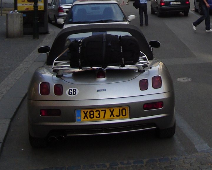 Pistonheads - The image depicts a busy city street where a silver sports car is parked on the side of the street alongside a black luggage rack full of luggage. The car is usually secured to the luggage rack, perhaps indicating that someone is about to embark on a long journey or has just arrived. In the background, there are other cars and pedestrians going about their day, contributing to the bustling atmosphere. All this takes place on a street lined with parked cars and building facades, typical of an urban environment.