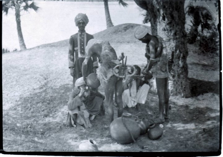 Pistonheads Identify - The image is a black and white photograph depicting a group of individuals in what appears to be a natural landscape. There are five people visible, with three standing and two sitting on the ground. They seem to be engaged in a gathering or a ritual. The person standing on the left appears to be in traditional attire, possibly indicating a cultural or historical context. They are holding what seems to be a staff or rod, which is commonly associated with certain ceremonies. In the center, another individual is bent over, interacting with an object on the ground. On the right, a person is looking closely at a smaller object, which could be a skull. In the background, there are palm trees, suggesting a warm climate environment. The overall setting, the attire of the people, and their actions hint at a cultural or traditional gathering.