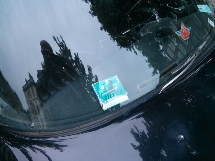 A rear view mirror on the side of a car - Pistonheads - The image captures the reflection of a person, partially obscured, on the rear window of a vehicle. The person's arm and legs are visible, but their face remains obscured due to the angle of the image. Positioned on the window is an item that appears to be a small, light-colored, square object with text on it, and there are also other stickers and marks on the glass. The setting suggests the person might be inside the vehicle, and the background shows a clear sky and an urban street scene.