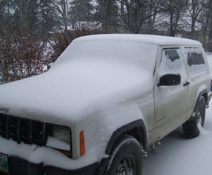 Jeep Cherokee 4.0 - Page 1 - General Gassing - PistonHeads - The image depicts a parked white jeep with a plow on the front facing the viewer. The vehicle is covered in a thick layer of fresh snow, which extends over its roof and onto its doors. The jeep is sitting on a road that appears to be in a neighborhood with a stylized roof visible in the background. There is a blurred text visible in the clear but overcast sky.