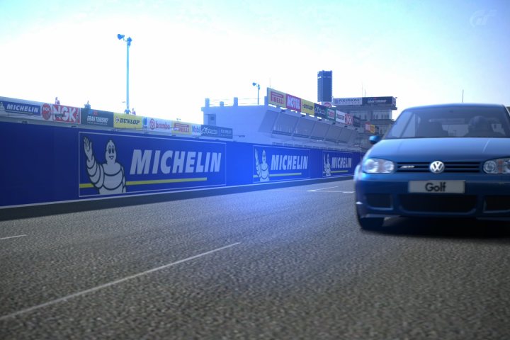 Gran Turismo 6 picture thread - Page 4 - Video Games - PistonHeads - This image depicts a blue VW Golf car driving on a track. The track is surrounded by a series of colorful sponsor banners, suggesting that it is a promotion for sponsors linked to the event or the circuit. In the distant background, the silhouette of an architectural structure, possibly stands or facilities, can be seen. The time of day may indicate either the early morning or late afternoon, given the lighting. The image seems to be a screenshot, possibly from a video game or a compilation of images showcasing the car.