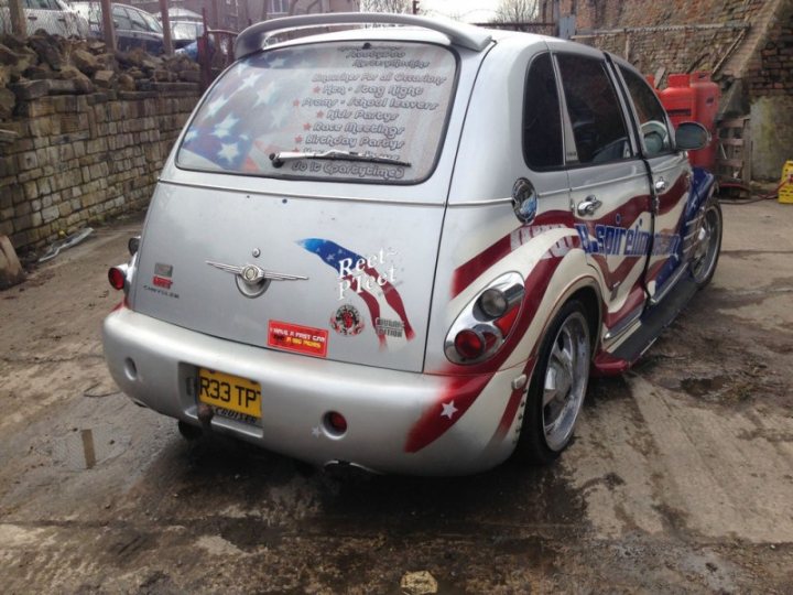 a bit council - Page 4 - The Lounge - PistonHeads - The image features a small, silver-colored car parked in a lot. The car is adorned with a noticeable paintjob of red, white, and blue colors that resemble the American flag, with the word "Pride" prominently displayed on the back. A license plate is visible, displaying "T25 TP." On the back windshield, there is a decal that reads "Reliever Concert's 2010," indicating the car may have been involved in a charity event. The surrounding area appears to be an industrial or yard setting with brick walls and equipment in the background.