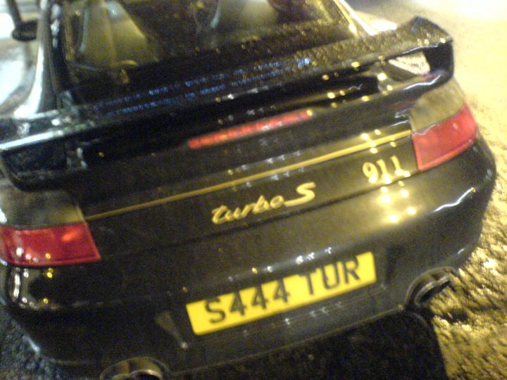 Pistonheads Misbadging - The image shows a black sports car parked on a street, as seen from a rear three-quarter angle. The car features a prominent rear spoiler and a brightly lit license plate that reads "S44 TUR." There are also tail lights visible. The car's glossy black surface reflects some lights, suggesting that it's either early evening or night time. The street itself appears to be wet, possibly from recent rain, enhancing the reflections on the car's body.