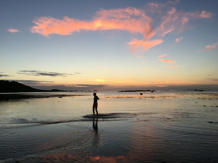 The 2016 summer and thunder thread - Page 25 - The Lounge - PistonHeads - The image depicts a serene beach scene at sunset. One person is seen standing in the shallow water at the water's edge, engaged in the act of photography or filming. The sky is painted with hues of orange and light blue, which cast beautiful colors onto the water's surface, enhancing the tranquil atmosphere. The horizon is visible and it looks tranquil with the calm sea meeting the sky. The combination of the exquisite sky colors, the silhouette of the person, and the tranquil sea create a captivating and peaceful scene.