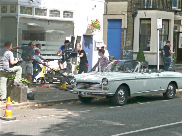 Anyone own a well known car from a tv series or film? - Page 4 - Classic Cars and Yesterday's Heroes - PistonHeads - The image captures a scene on a city street where an old-fashioned blue Mercedes-Benz car is parked near the curb. A young man in a light-colored shirt, dark pants, and sunglasses is seen seemingly directing traffic while holding what appears to be camer equipment. There are numerous people in various states of action in the background, some near a building entrance and others ridding a powered cart. The presence of the car and the bustling activity suggest a day in a busy city neighborhood.