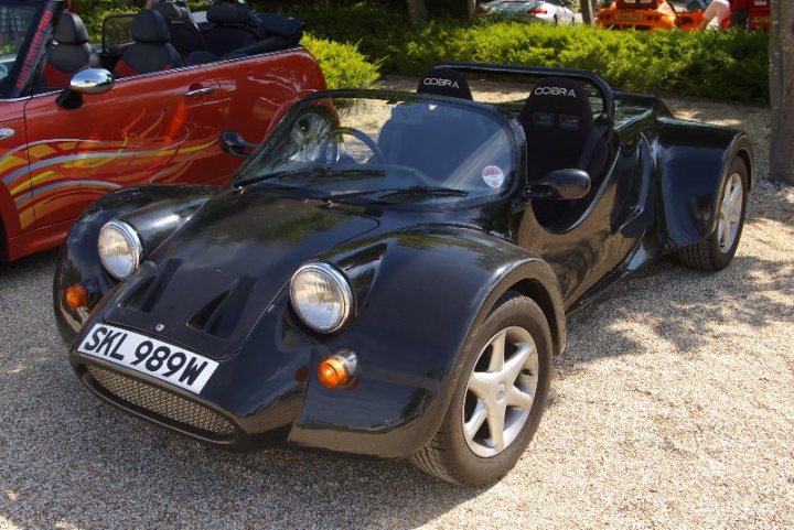 Pistonheads - The image features two vehicles parked in a lot, a car with a distinctive red flame design on the side and a smaller sports car in dark colors. The sports car is the one more prominently in the foreground, with a clean and well-maintained appearance, indicating it might be on display. The car has a sleek body with large, round headlights and a visor. It's difficult to make out specific details due to the distance and angle of the photograph, but it appears to be an open-air model, possibly a single-seater or a two-seater sports car. There are no visible texts or distinctive emblems on the vehicles that are recognizable. The background shows the corner of a parking lot with some trees and greenery, and a small water feature is also barely visible to the right of the vehicles, suggesting the area might be designed to be more aesthetically pleasing.