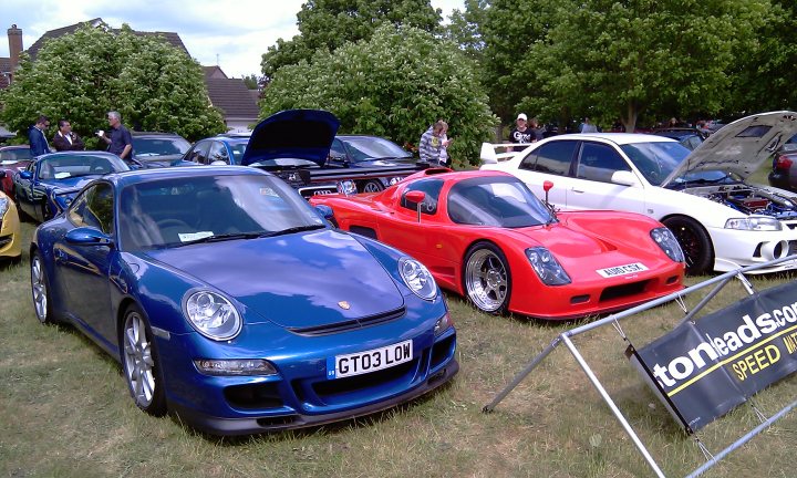 Cars On The Green - Photo Thread - Page 1 - East Anglia - PistonHeads - The image captures an outdoor scene at a car meet where various cars are on display. The cars, painted in different colors like blue, red, and white, are parked on grass behind a fence, suggesting a casual and open environment. Some cars are modified with visible additions or aftermarket parts. The scene conveys a sense of camaraderie and shared interest among car enthusiasts.